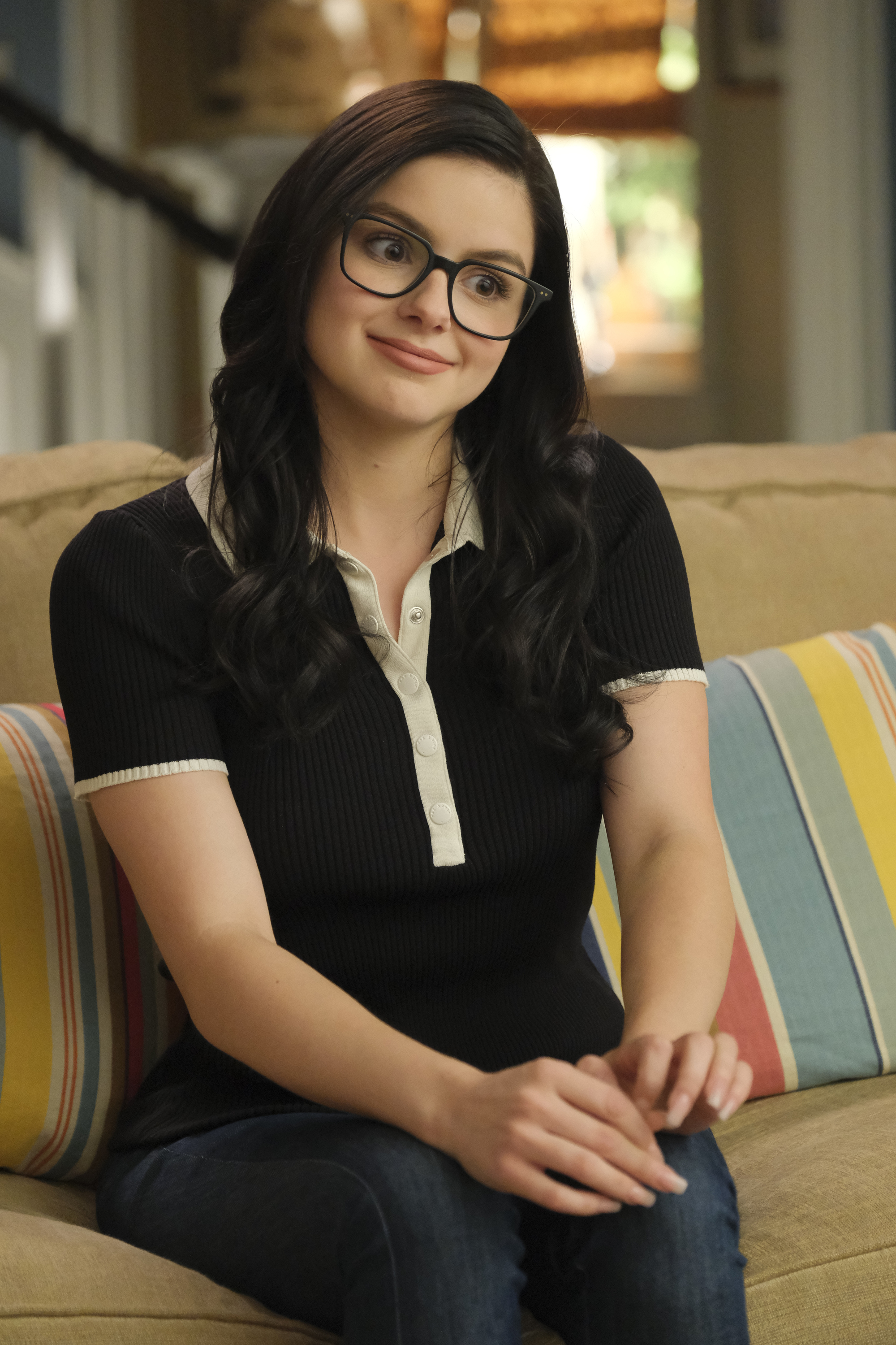 A woman with long dark hair and glasses sits on a couch, wearing a black short-sleeve collared shirt and jeans