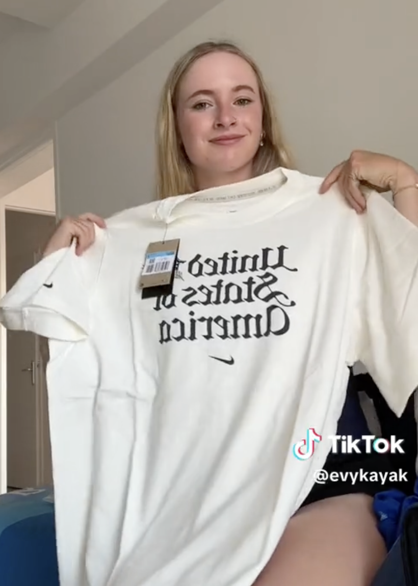 Person holding a white t-shirt with &quot;United States of America&quot; text. TikTok username &quot;@evykayak&quot; is visible