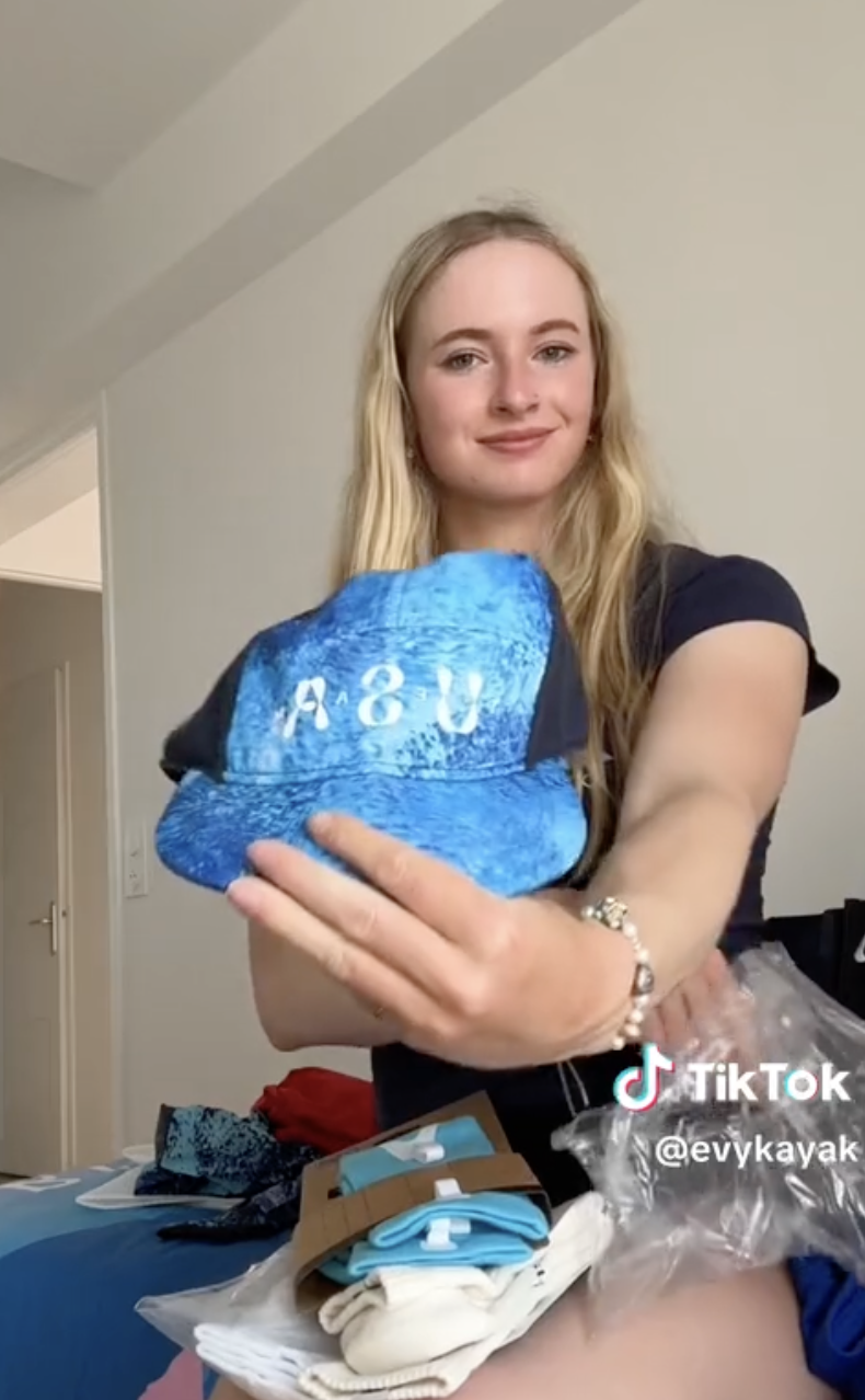 Evvy Kayak, sitting, holds up a blue A&amp;amp;V hat in a room. The TikTok username @evvykayak is visible