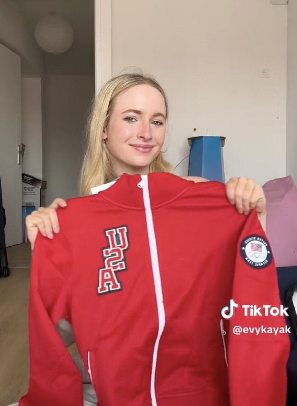 Unidentified person smiling and holding a red sports jacket with &quot;USA&quot; and Olympic patches. TikTok username &quot;evykayak&quot; visible on the image