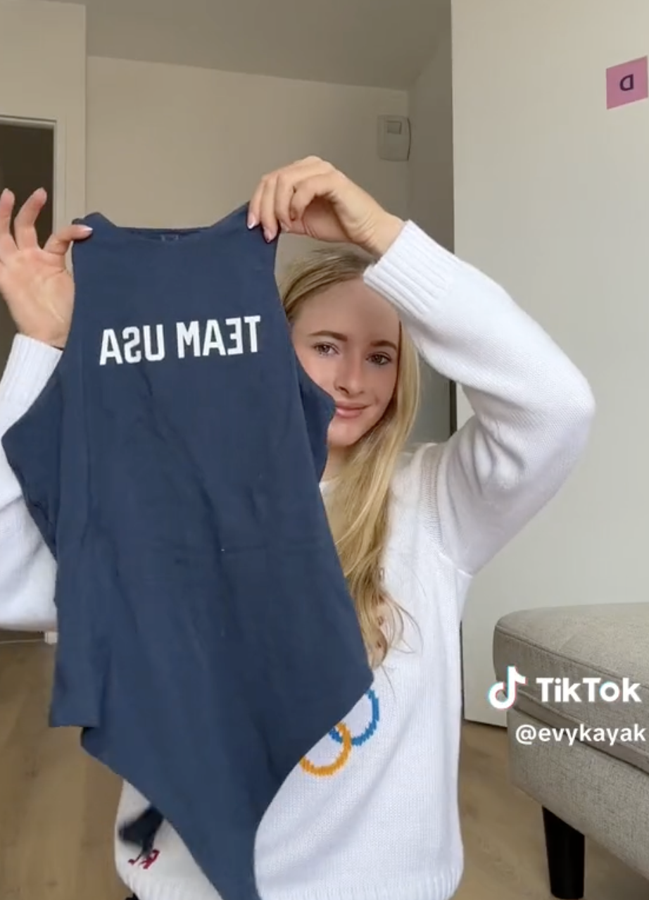 Evy Poumpouras holds up a &quot;Team USA&quot; tank top in a TikTok video by @evykayak. She is wearing a white sweater and is indoors