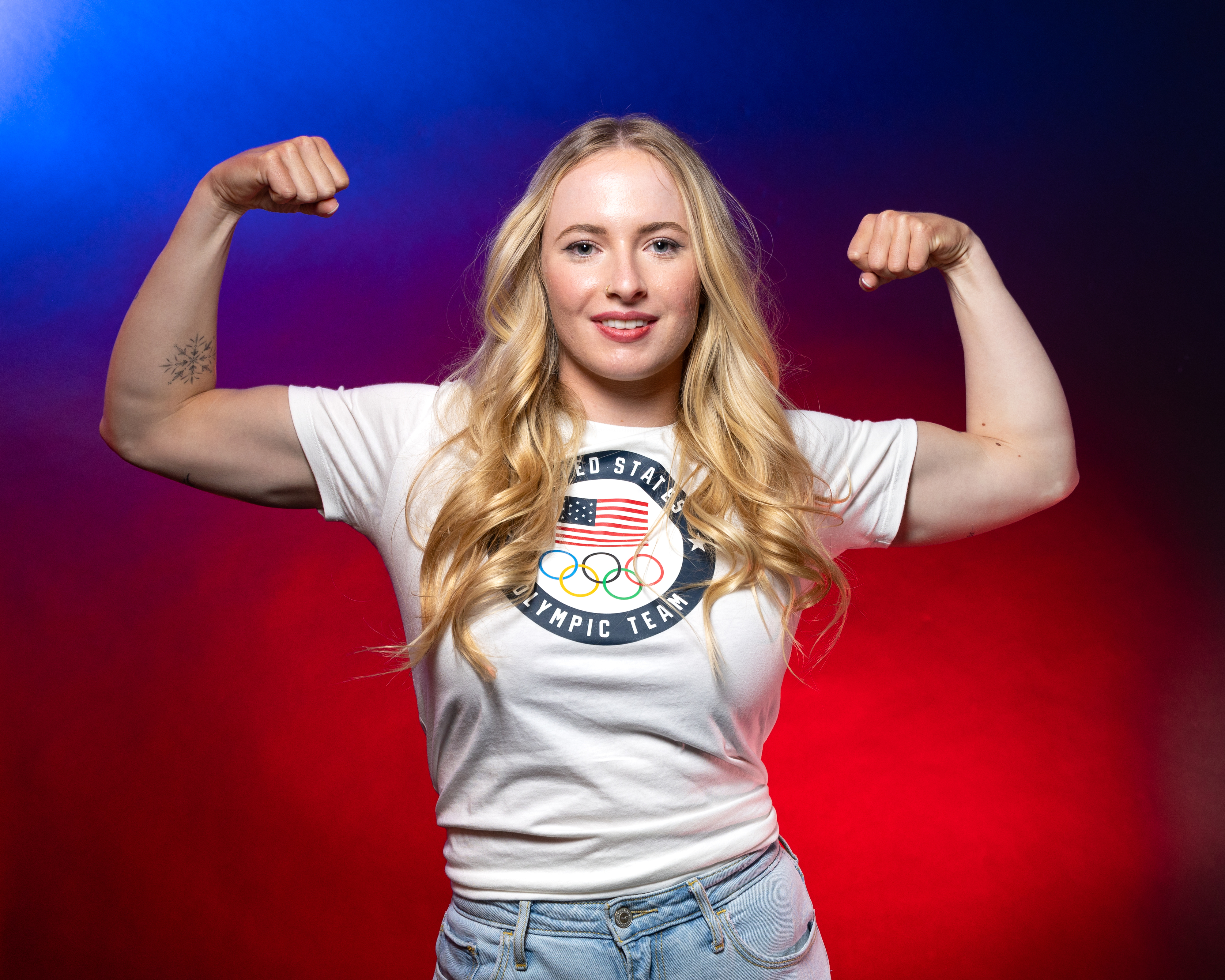 Athlete flexing biceps while wearing a USA Olympic Team t-shirt and jeans, smiling confidently at the camera