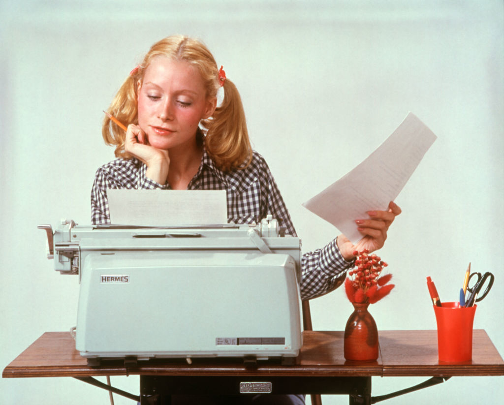 Shirley Jones types on a Hermes typewriter, holding a paper with one hand while resting her chin in the other. A red vase with flowers and desk organizer are beside her