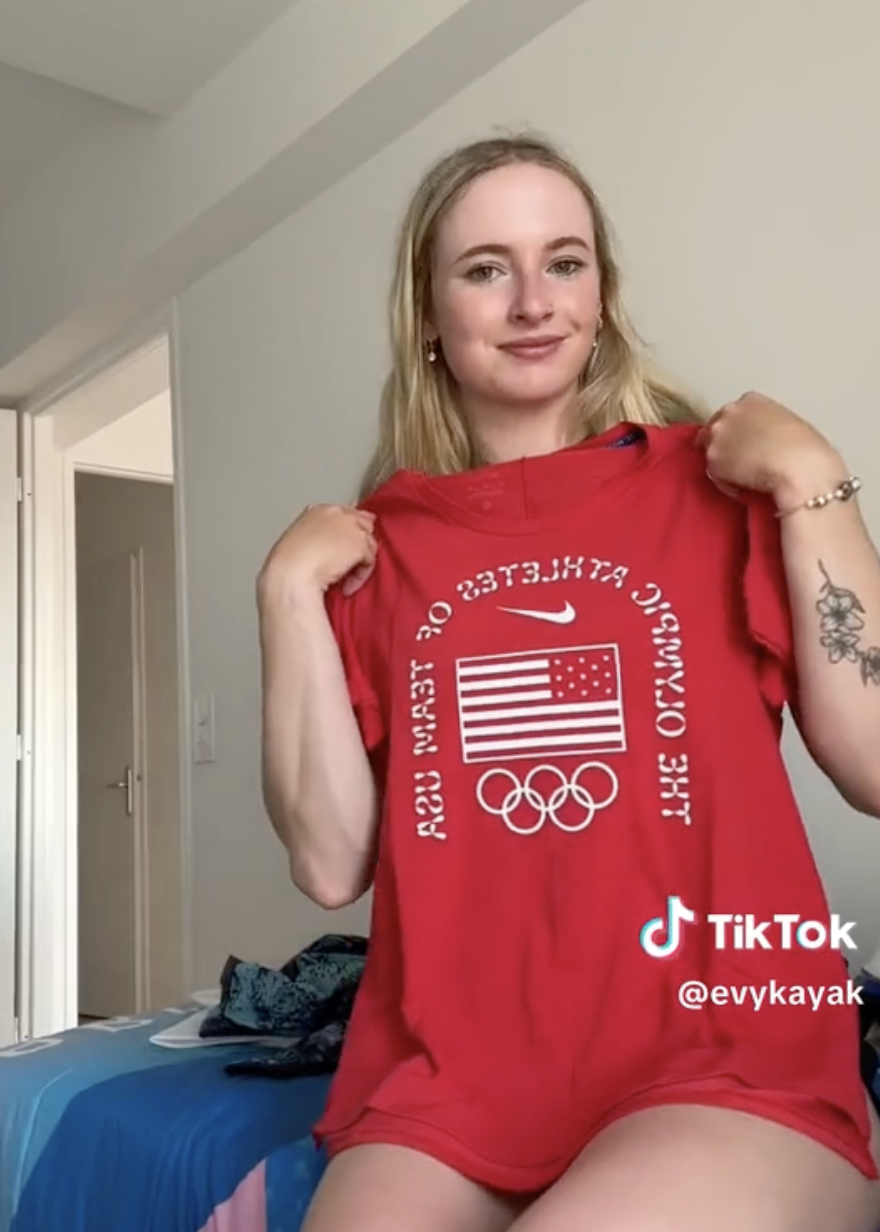 Evy Kayak is sitting and holding up a red Team USA Olympic t-shirt, smiling at the camera. The image features a TikTok handle @evykayak