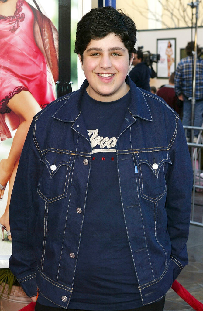 Josh Peck smiles while wearing a denim jacket and a graphic t-shirt at a red carpet event. Poster of a woman in the background