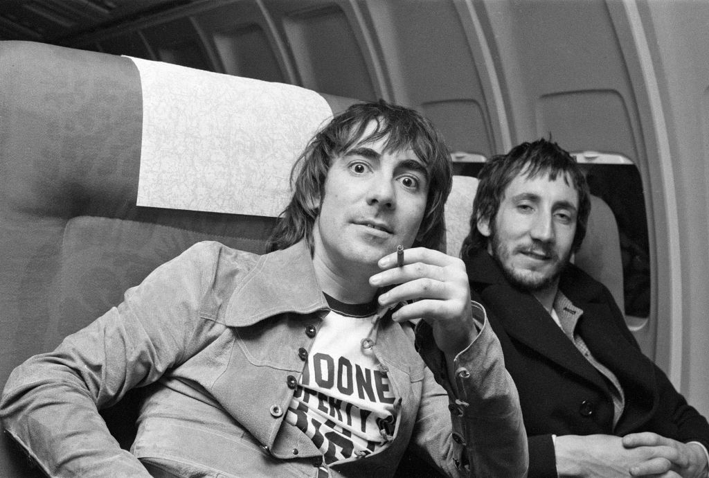 Keith Moon and Pete Townshend sit in an airplane, with Keith holding a cigarette and wearing a T-shirt, and Pete in a dark coat, both looking at the camera