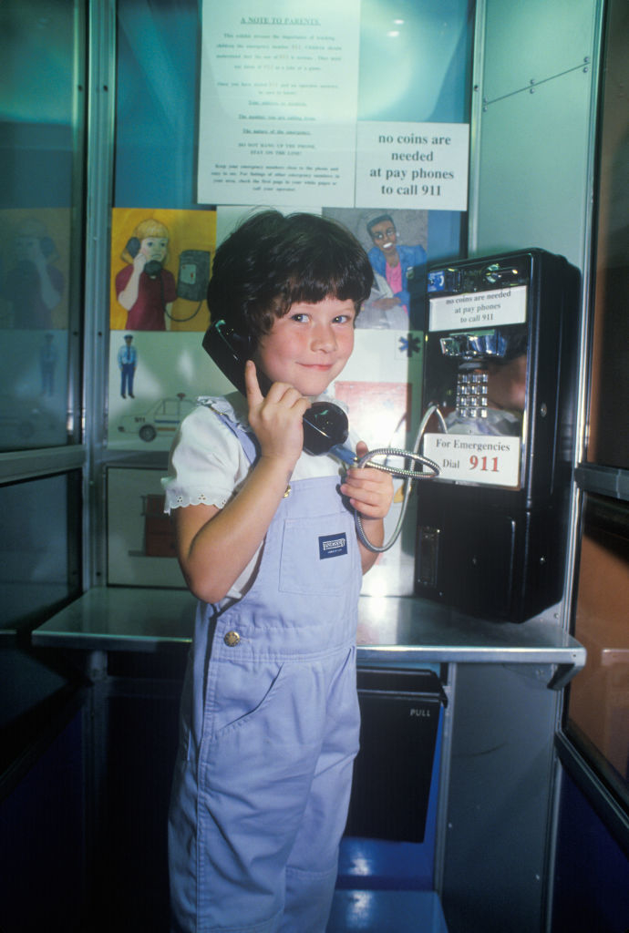 A young child in a vintage phone booth holding a phone receiver to their ear, smiling. Signs on the booth mention that no coins are needed to call 911