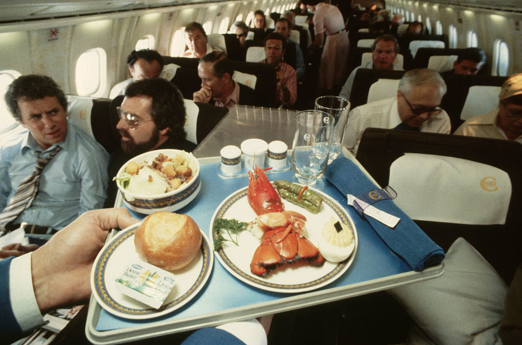Passengers seated on an airplane look ahead while a flight attendant holds a tray with lobster, a roll, and side dishes in front of them