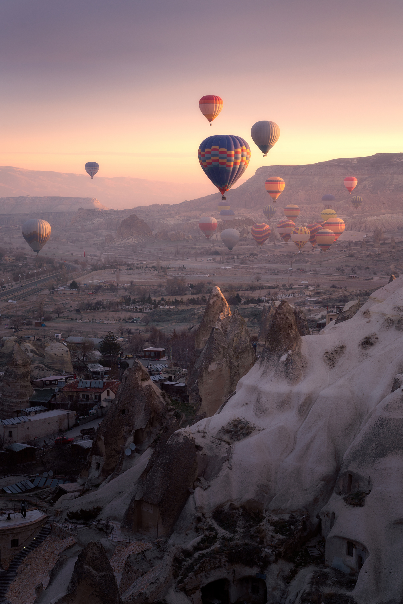 Hot air balloons float over a rocky landscape with a small town below during a sunrise or sunset