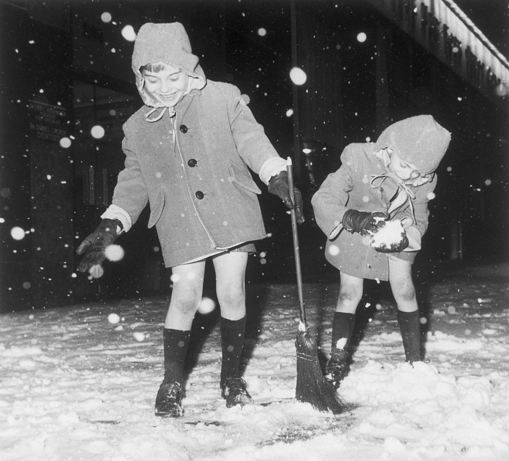 Two children, wearing coats and winter hats, play in the snow. One child sweeps snow with a broom, while the other gathers snow in their hands