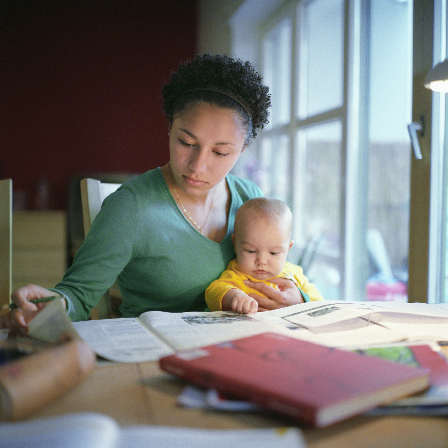 A woman sits at a table reading a book while holding a baby on her lap. The table is cluttered with books and papers