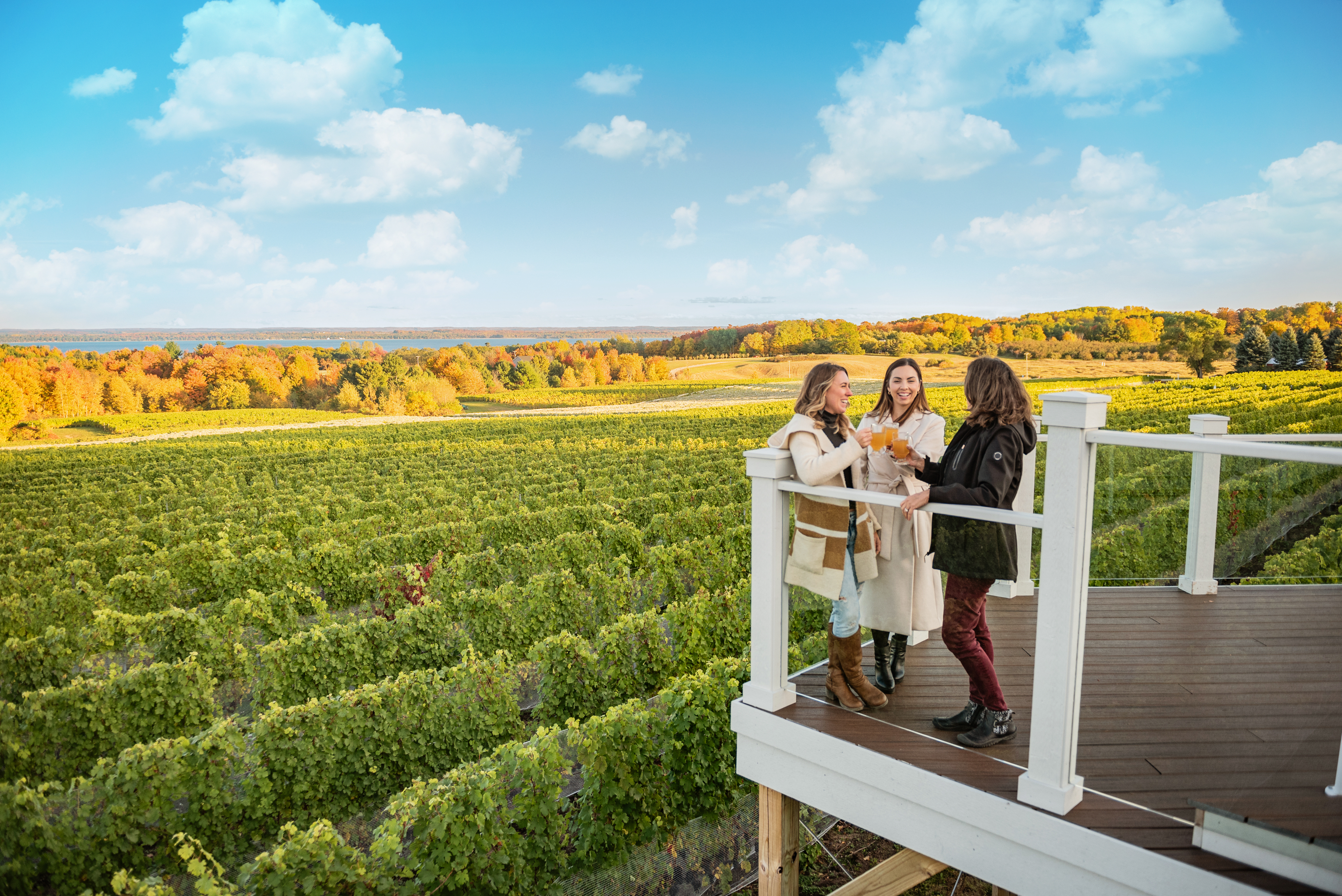 Three women enjoy wine and conversation on a deck overlooking a vineyard with autumn foliage