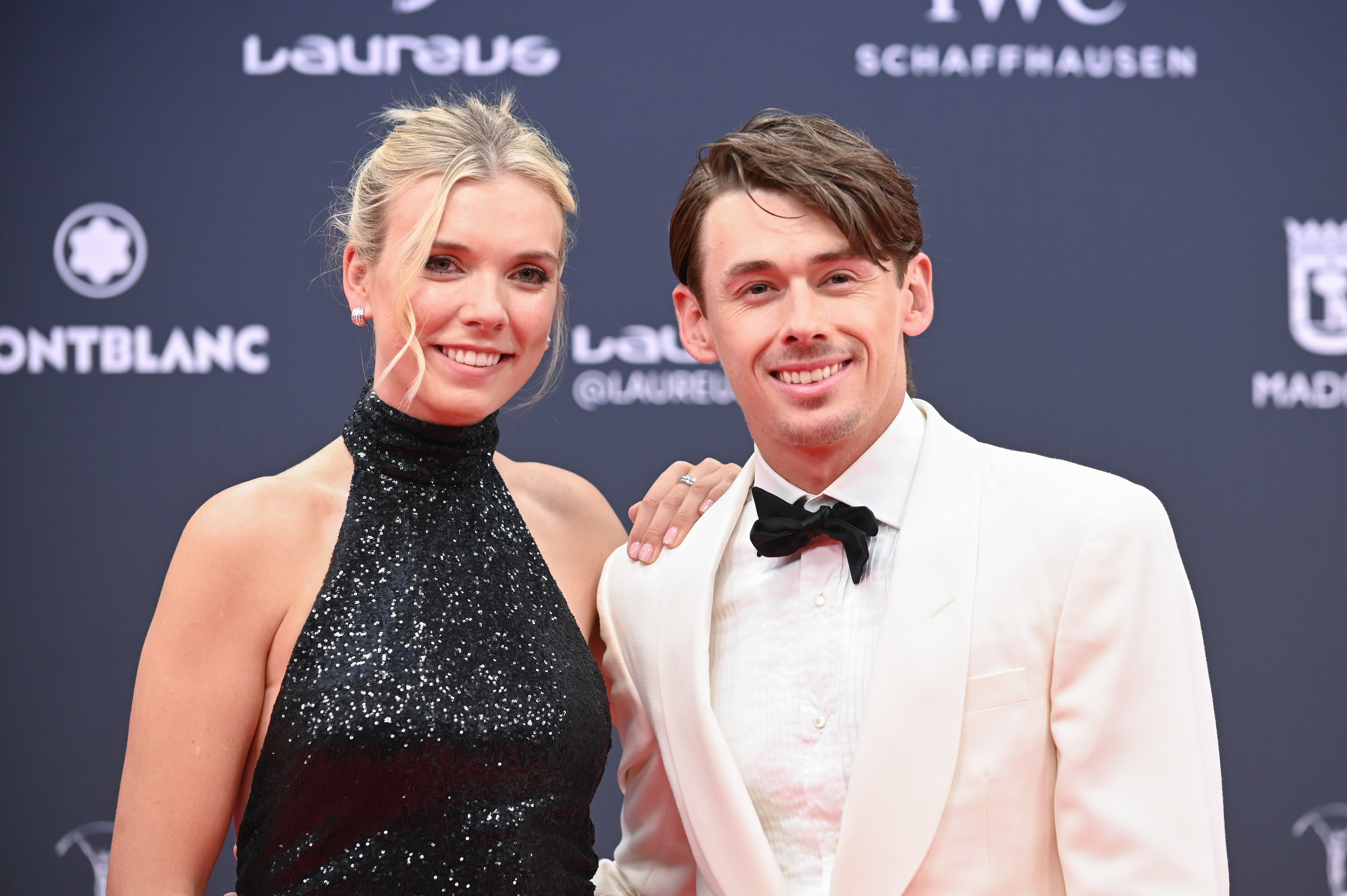 Two people pose on the red carpet. The woman wears a sleeveless, high-neck sparkling dress; the man wears a white suit with a black bow tie. Names not provided