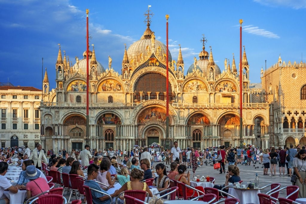 A crowded scene in Saint Mark's Square, Venice, with tourists seated at a café and St. Mark's Basilica in the background