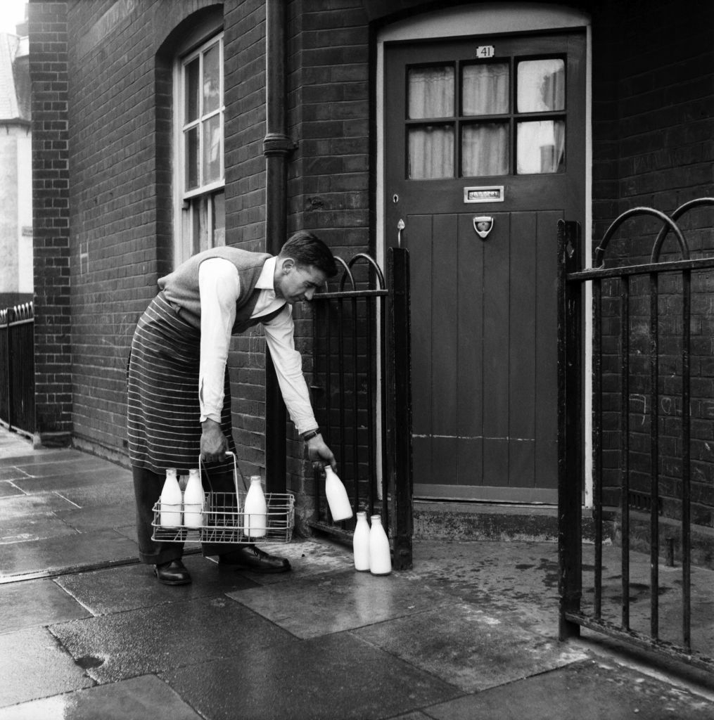 A milkman in a striped apron and shirt is delivering glass milk bottles to a doorstep in front of a brick building