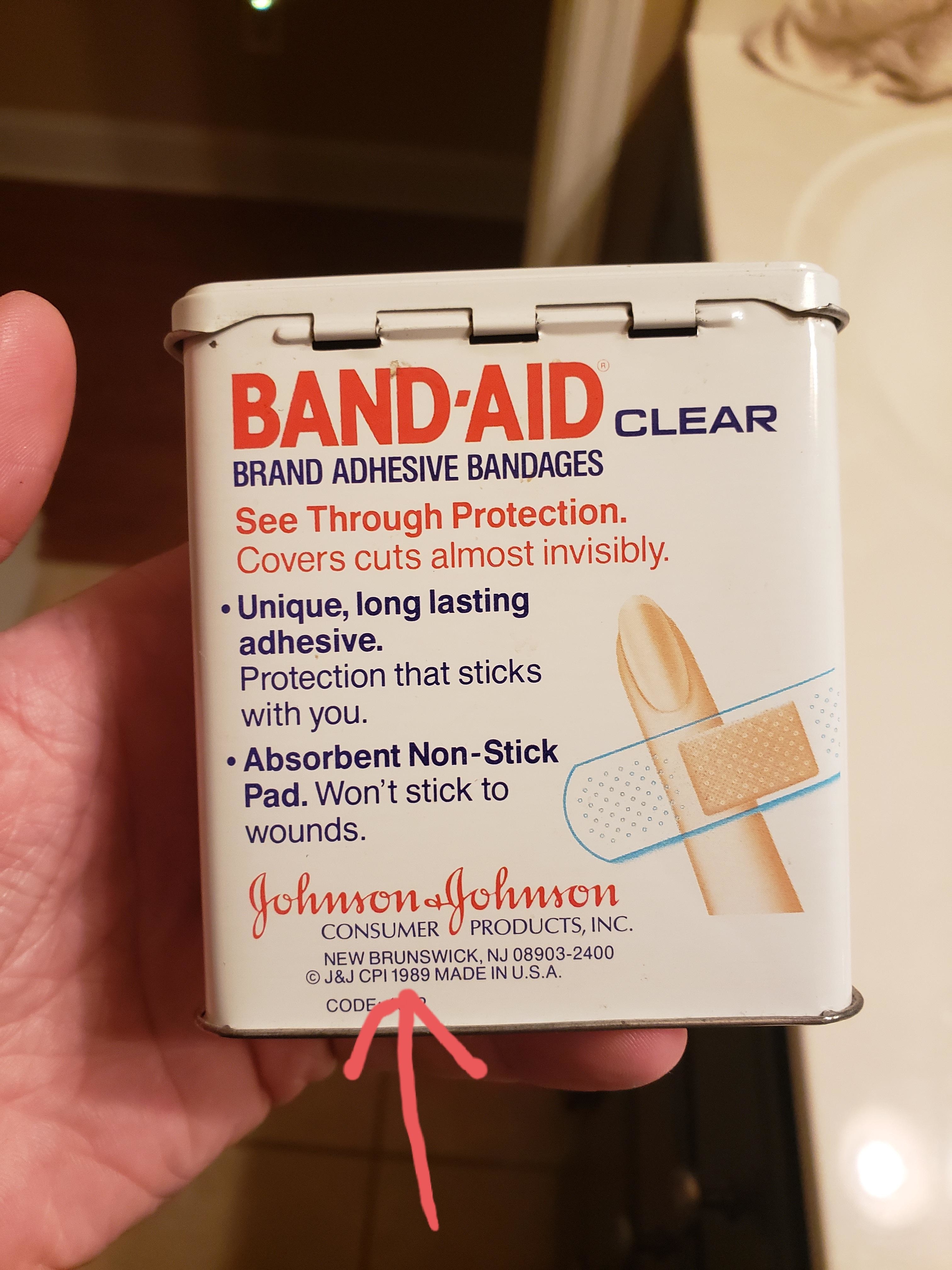 A hand holding a box of BAND-AID brand adhesive bandages with text describing its benefits and manufacturer information