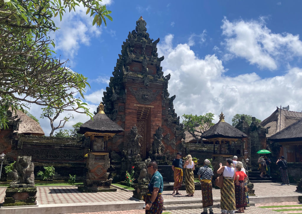 People are visiting a traditional Balinese temple with intricate carvings and statues. Some visitors are taking photos, and others are walking around the temple grounds