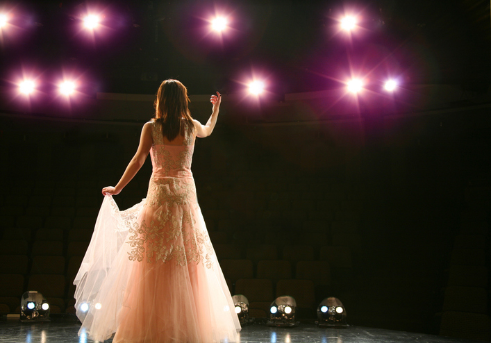 Woman on stage in an elegant dress, holding the hem of her skirt, under five bright spotlights, facing away from the camera towards an empty audience