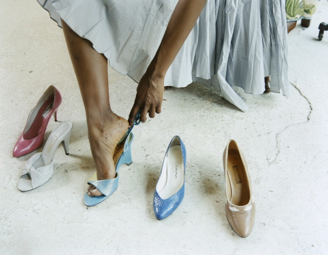 A person wearing a long skirt tries on a blue high-heeled shoe, surrounded by other high-heeled shoes in various styles on the floor