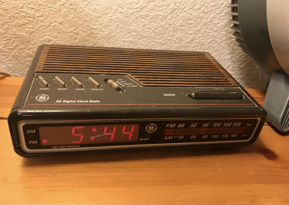 A vintage General Electric digital clock radio displays the time 5:44 PM, set on a wooden surface near a corner of a room