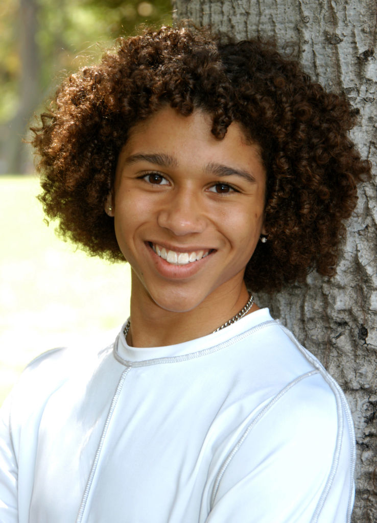 A young person with curly hair, wearing a simple white shirt, smiles while leaning against a tree