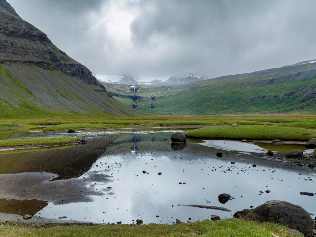 A serene landscape featuring a calm pond with reflections of surrounding mountains and a cascading waterfall in the distance