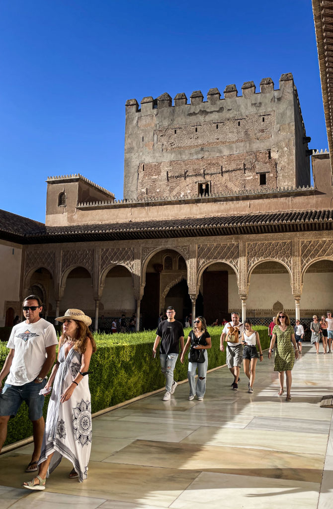People walk through a historic courtyard. There is a large, ancient stone building in the background with detailed architectural features
