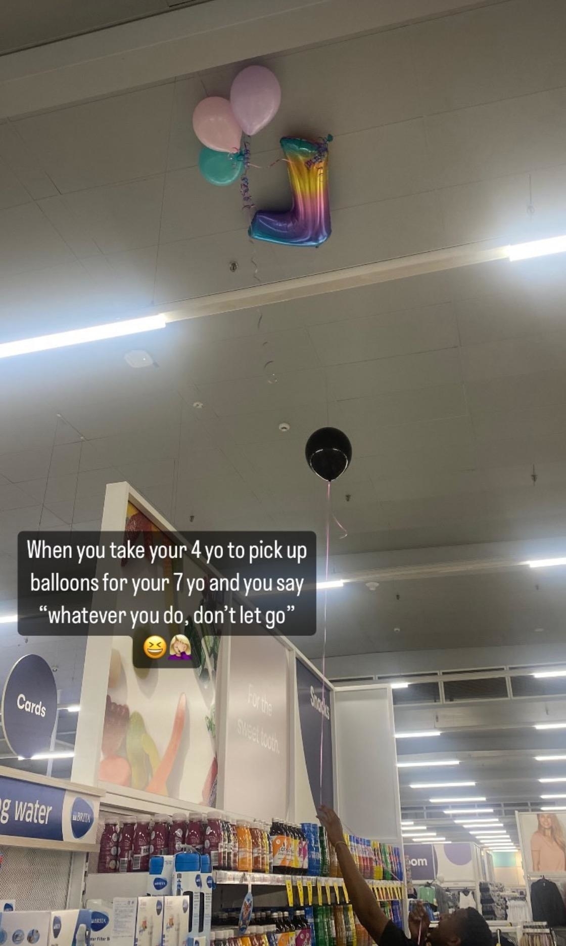 A person uses a long stick to retrieve balloons from a grocery store ceiling. Overlaid text reads, "When you take your 4 yo to pick up balloons for your 7 yo and you say 'whatever you do, don't let go'"