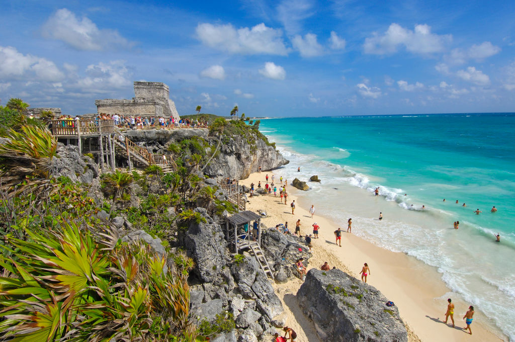Scenic beach with people swimming and sunbathing near ancient ruins on a rocky coastline in Tulum, Mexico