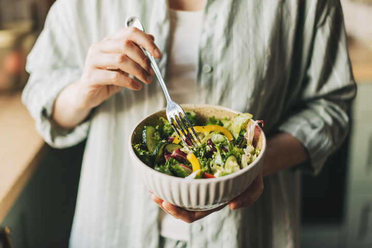 Person holding a bowl of salad with various vegetables, using a fork to eat