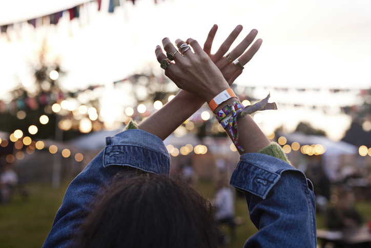 Person at an outdoor event, seen from behind, raising hands adorned with rings and festival wristbands, with string lights and banners hanging in the background