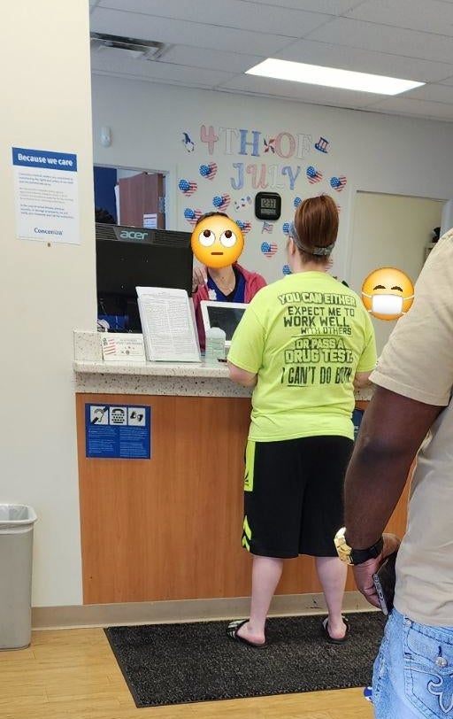 Two people stand in line at a counter inside a room. One wears a beige shirt and shorts, the other a bright green shirt with text. Faces are covered by emojis