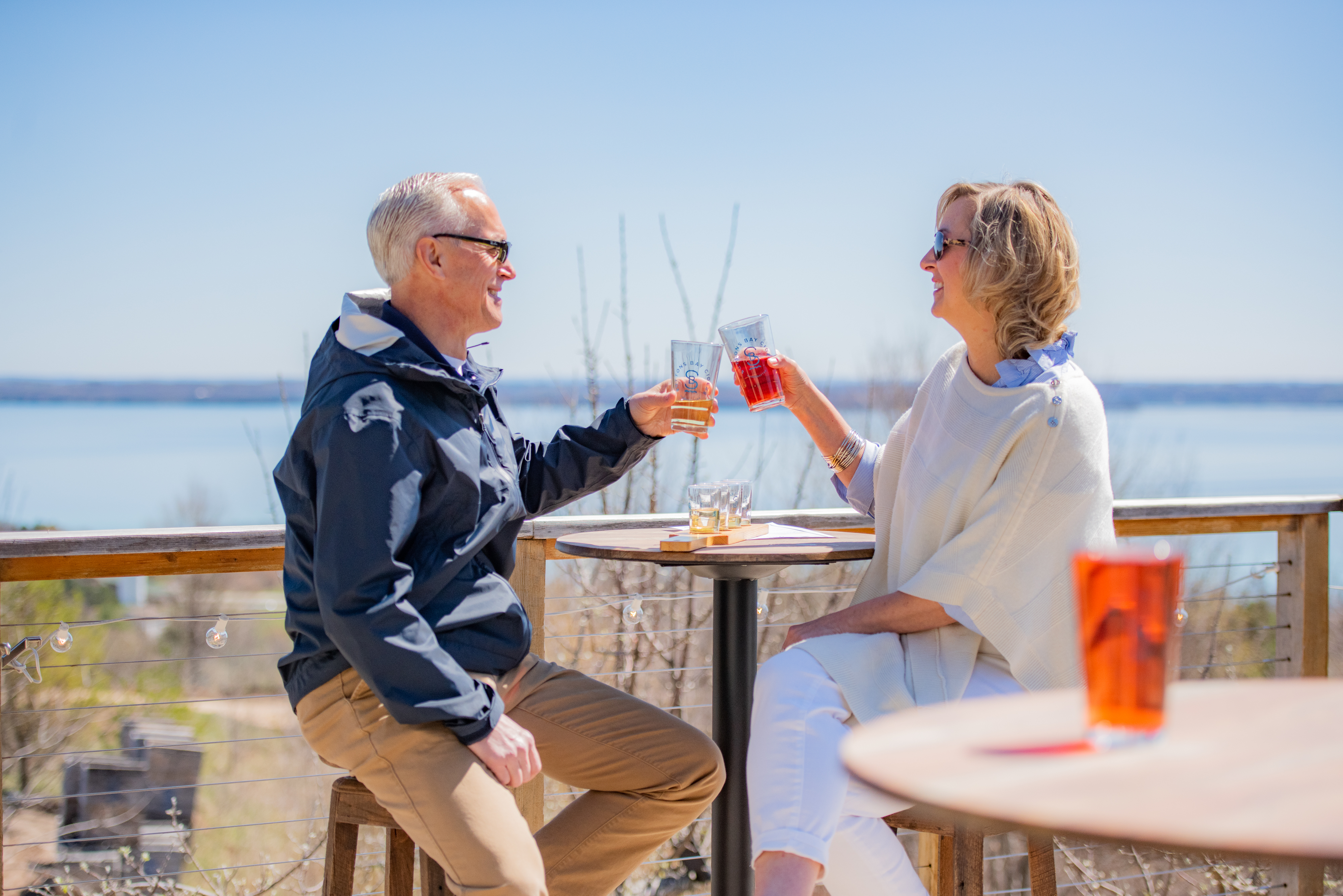 Two people clink glasses at an outdoor setting with a scenic backdrop. They are casually dressed, smiling, and appear to be enjoying drinks