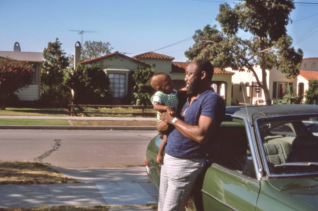 Man holding a baby while standing next to a car in a residential neighborhood
