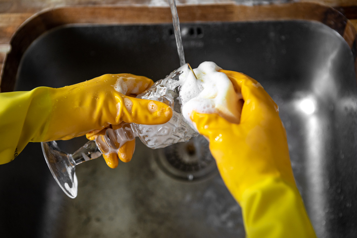 Hands wearing yellow gloves washing a glass under running water in a sink