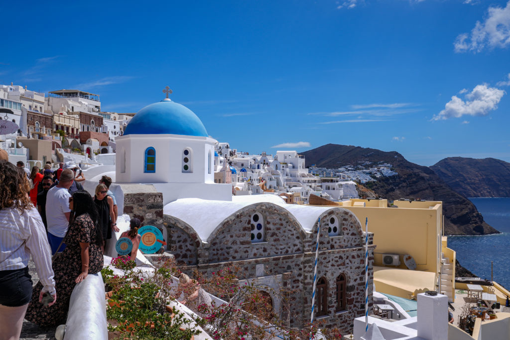 People explore white-washed buildings and a blue-domed church in Santorini, Greece, with a cliffside view of the sea in the background