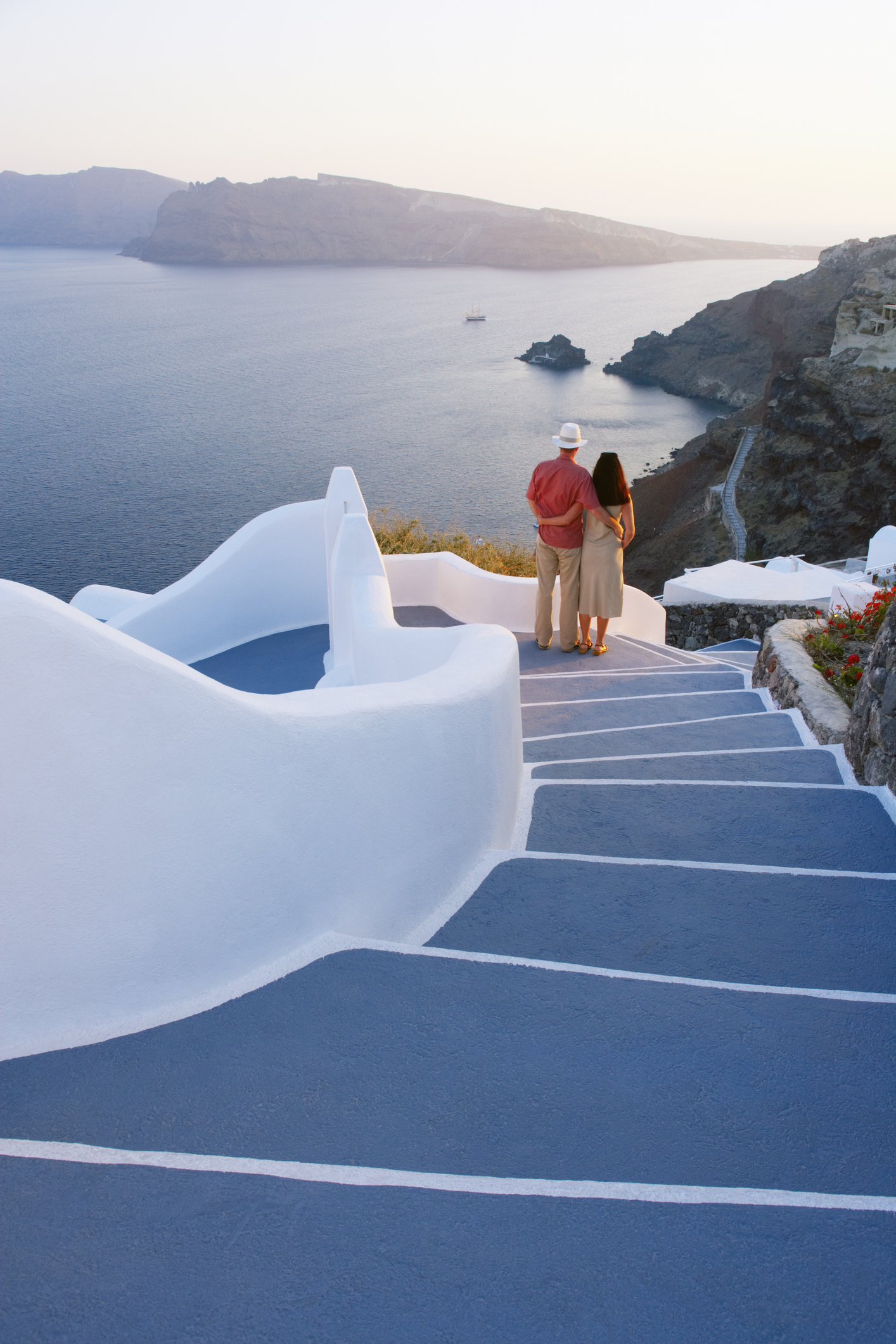 A couple stands at the bottom of white and blue stairs overlooking a scenic ocean view and coastline in Santorini. The man wears a red shirt and white hat; the woman a beige dress