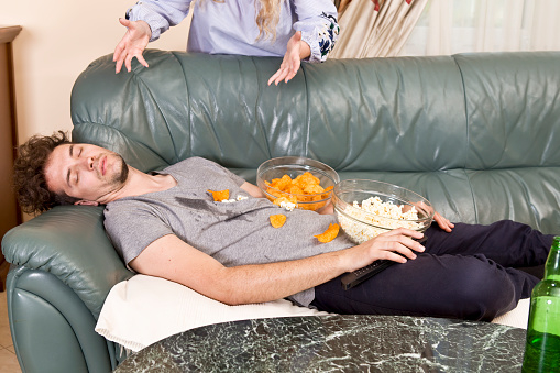 A man lies asleep on a couch with bowls of popcorn and chips on his chest