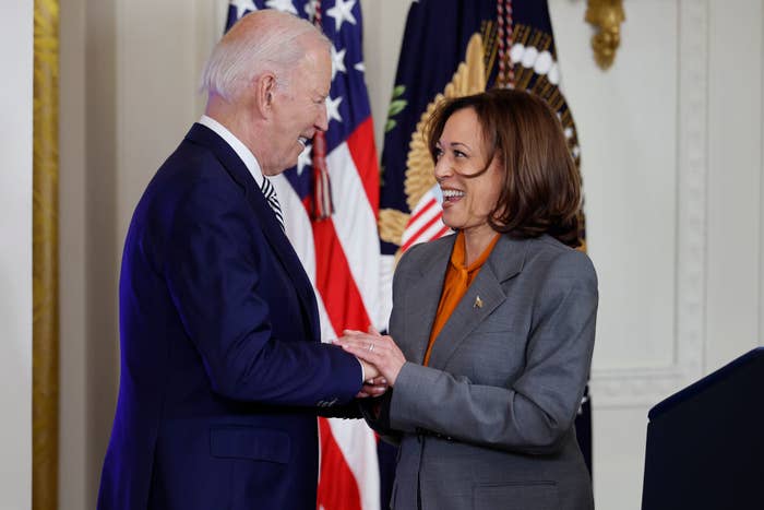 President Joe Biden and Vice President Kamala Harris share a handshake and smile during an event with U.S. flags and the presidential seal in the background