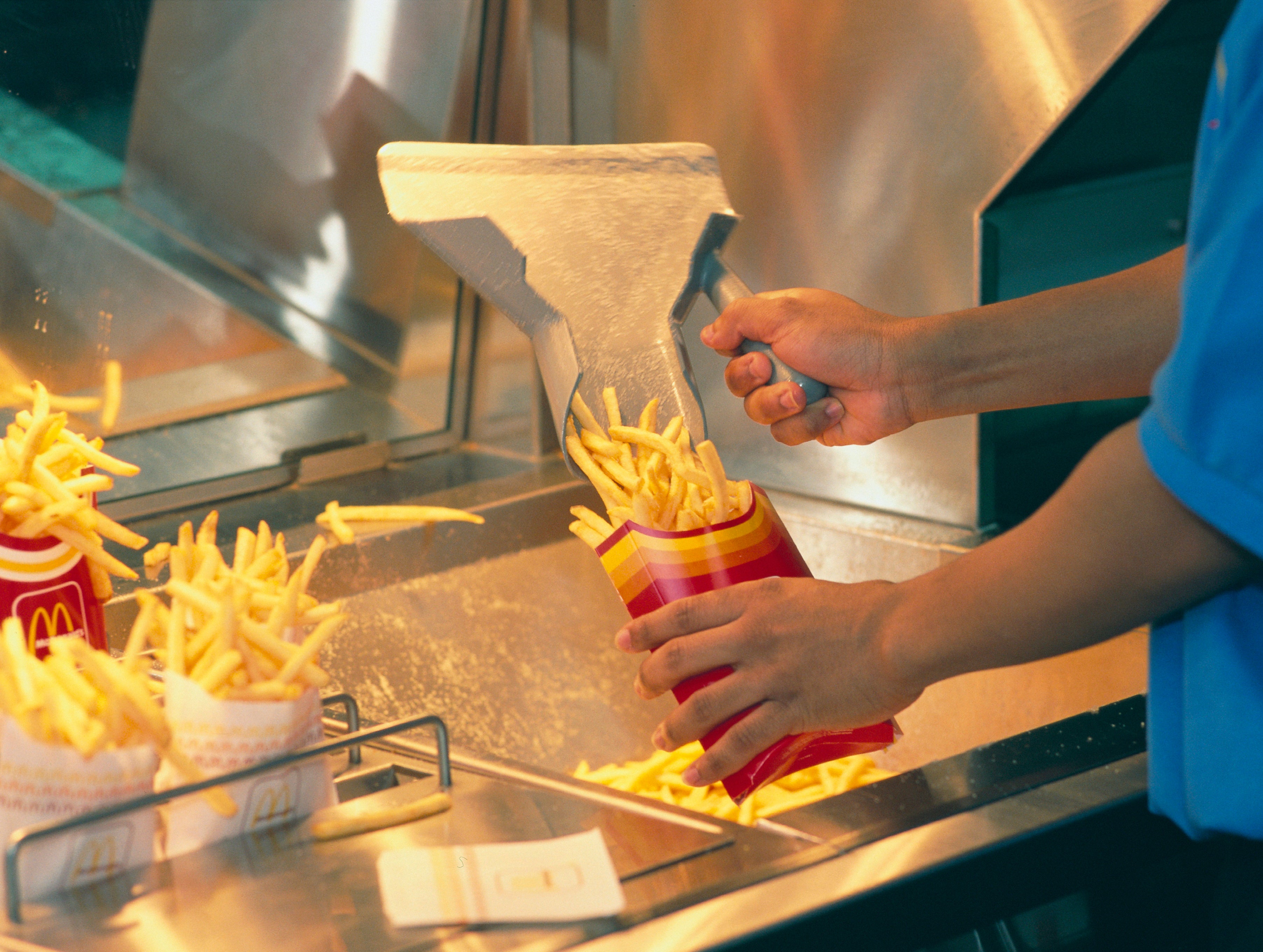 A worker at McDonald's fills a red and yellow fries container with freshly cooked French fries using a metal scoop