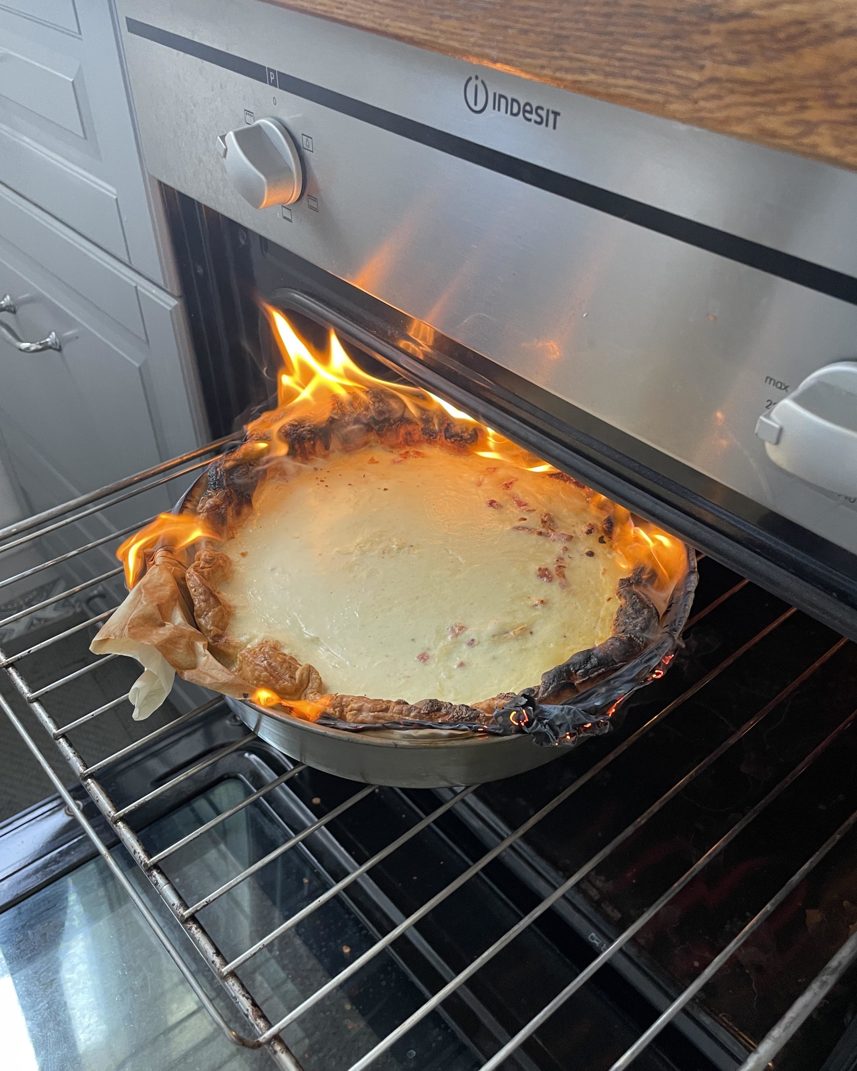 A burning pie with flames on the edges sits in an open oven, with smoke rising from it. The kitchen cabinets and oven controls are visible