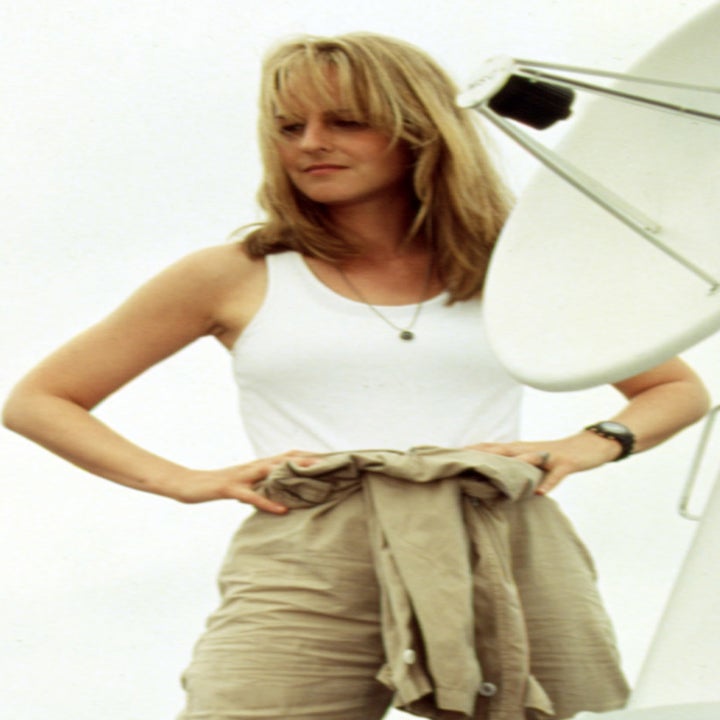 Helen Hunt stands confidently next to a satellite dish, dressed in a white tank top and light-colored cargo pants, in a scene from a movie