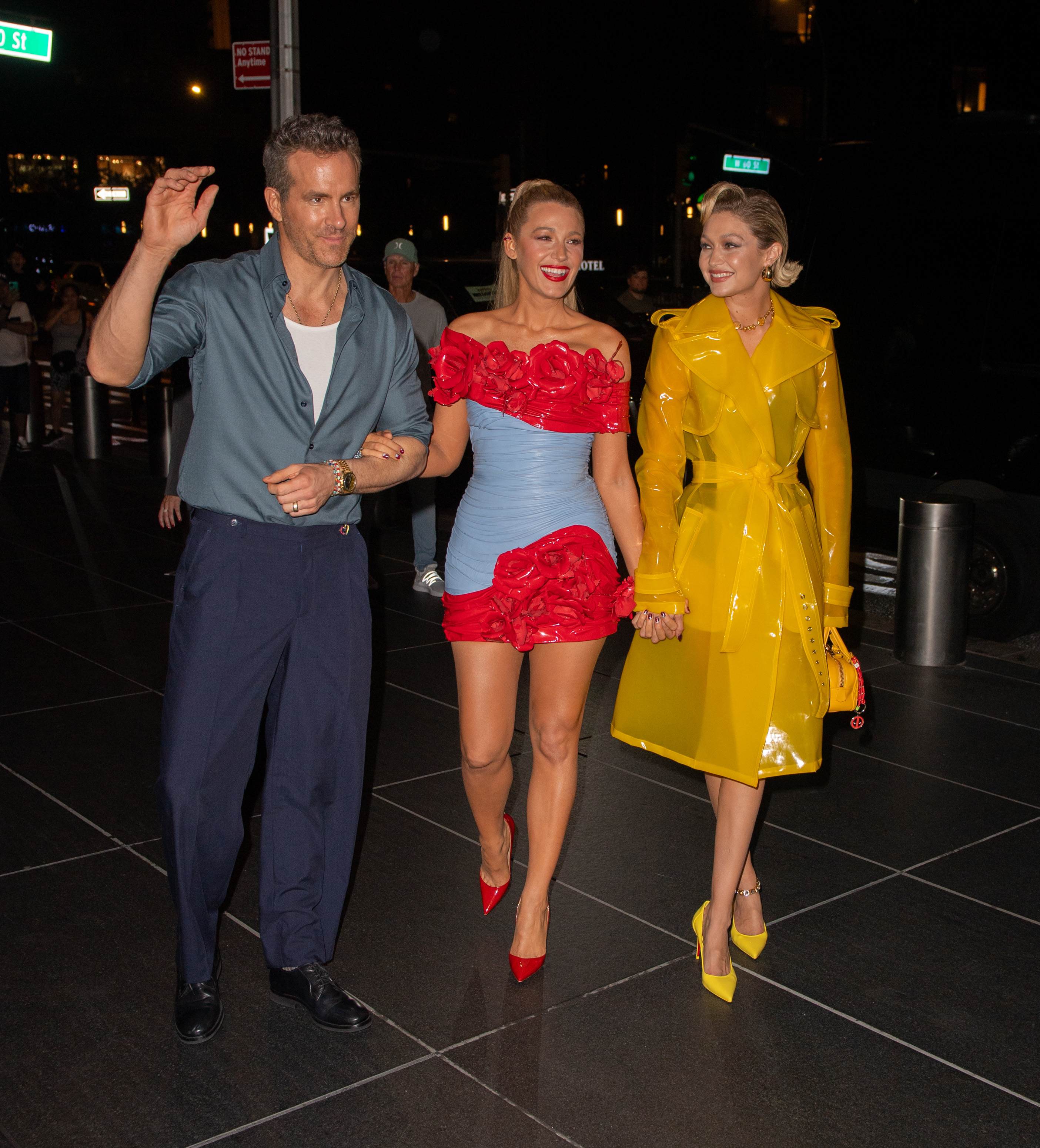 Ryan Reynolds, Blake Lively, and Gigi Hadid stand together outdoors. Ryan waves, Blake wears a short, strapless dress with floral details, and Gigi wears a yellow trench coat