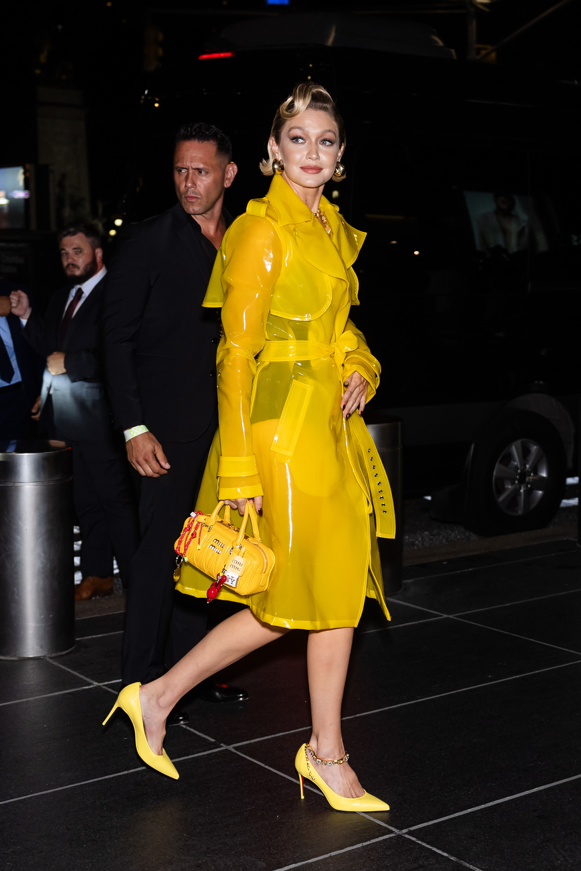 Gigi Hadid walks in a stylish clear yellow trench coat with matching yellow heels and purse, accompanied by a security guard