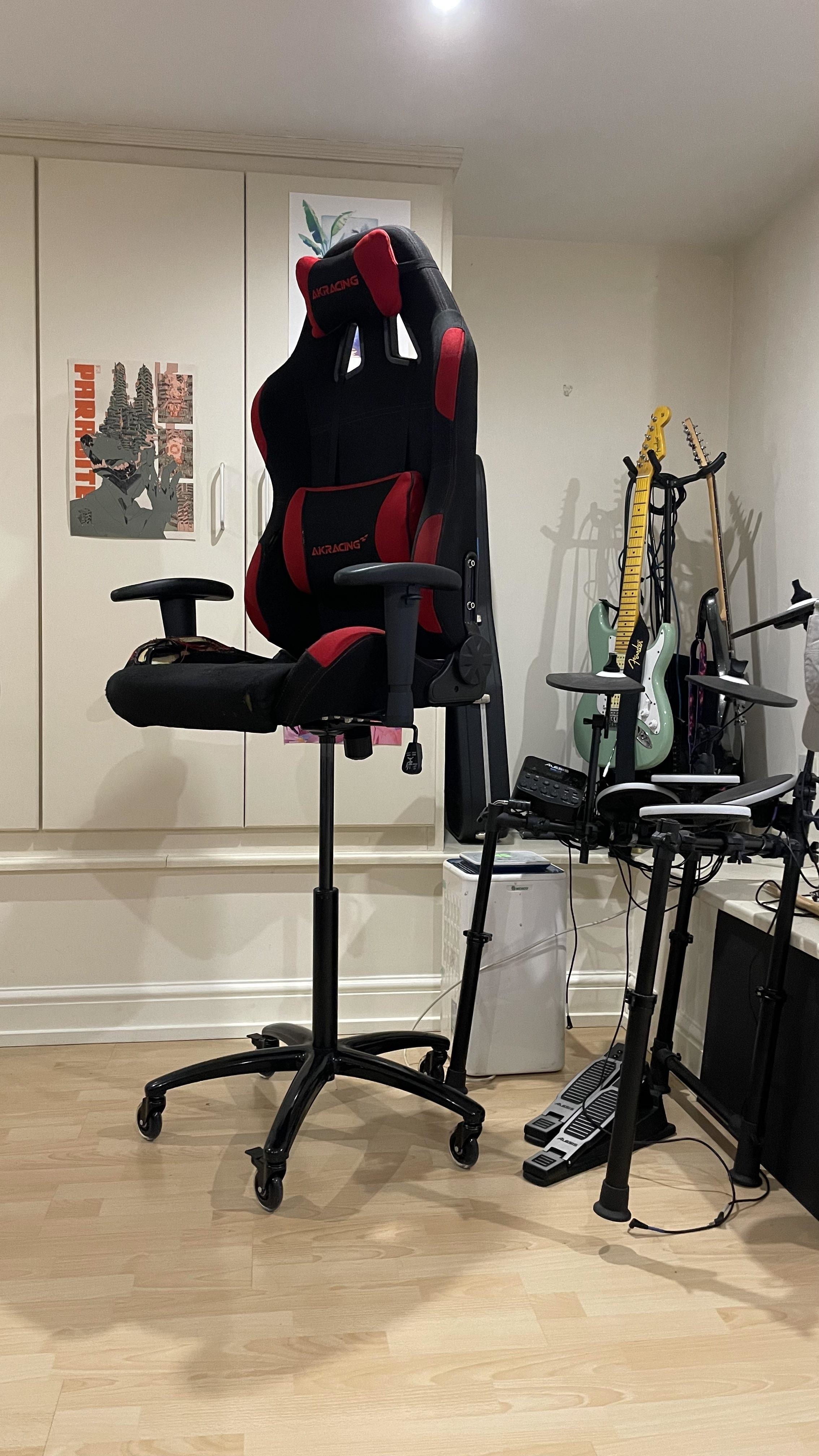 Gamer setup with ergonomic racing chair, electric guitar, bass guitar, drum set, and assorted gaming gear in a small, tidy room