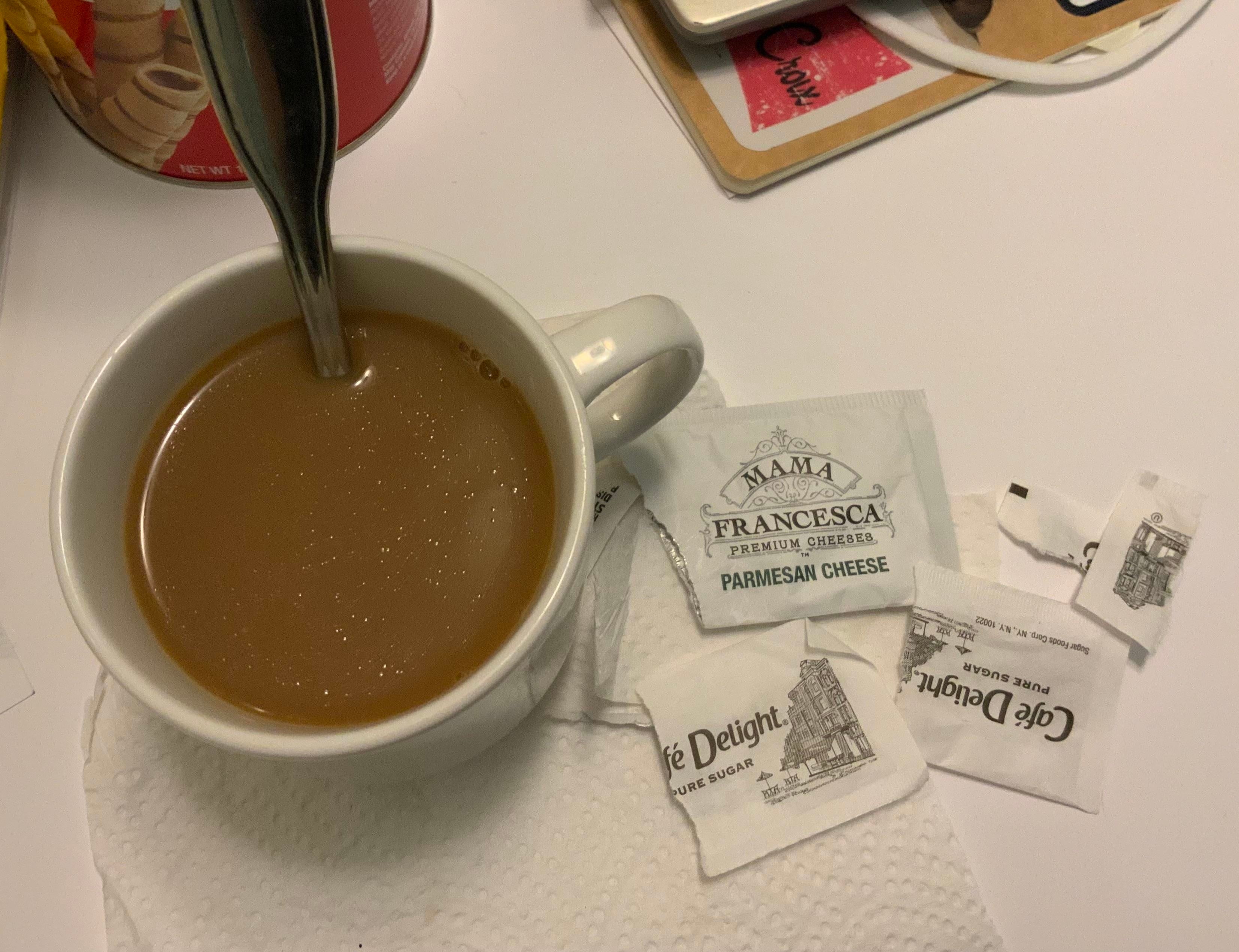 A cup of coffee with a spoon sits on a napkin next to several packets of grated Parmesan cheese