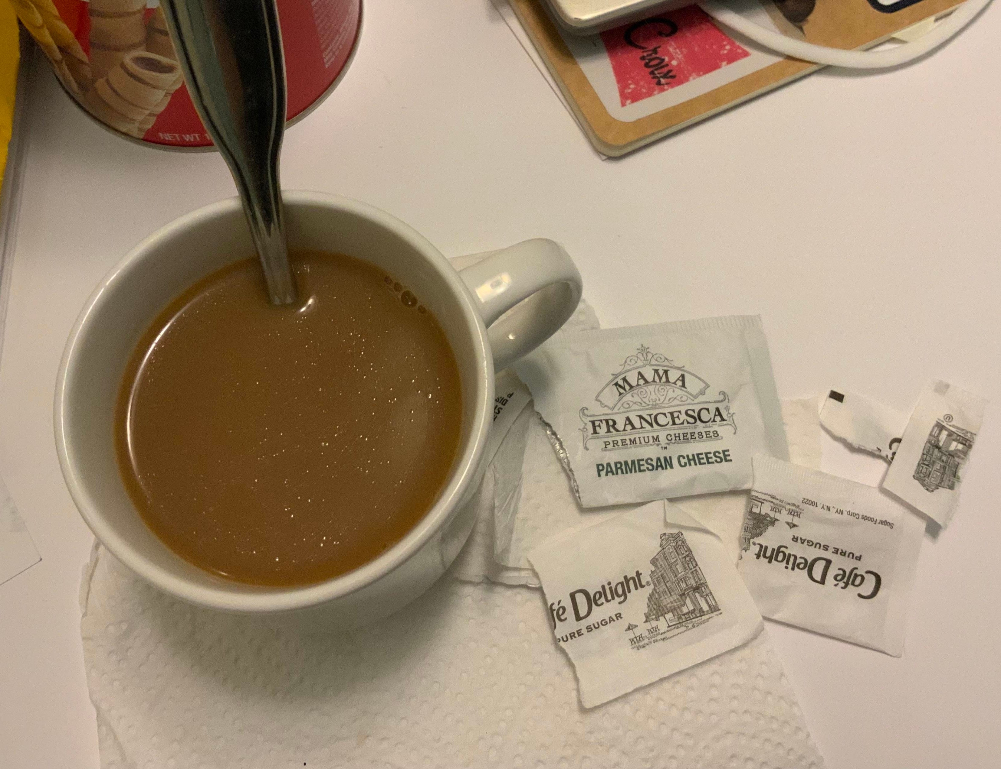 A cup of coffee with a spoon sits on a napkin next to several packets of grated Parmesan cheese