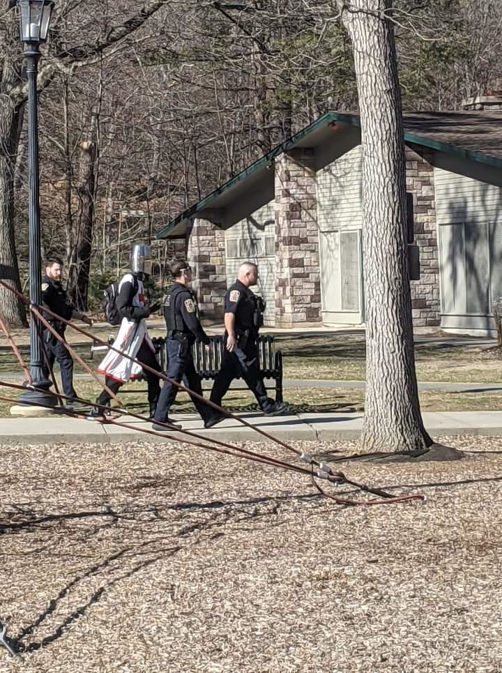 Three police officers escort a person wearing a Templar knight costume through a park past a stone building on a sunny day