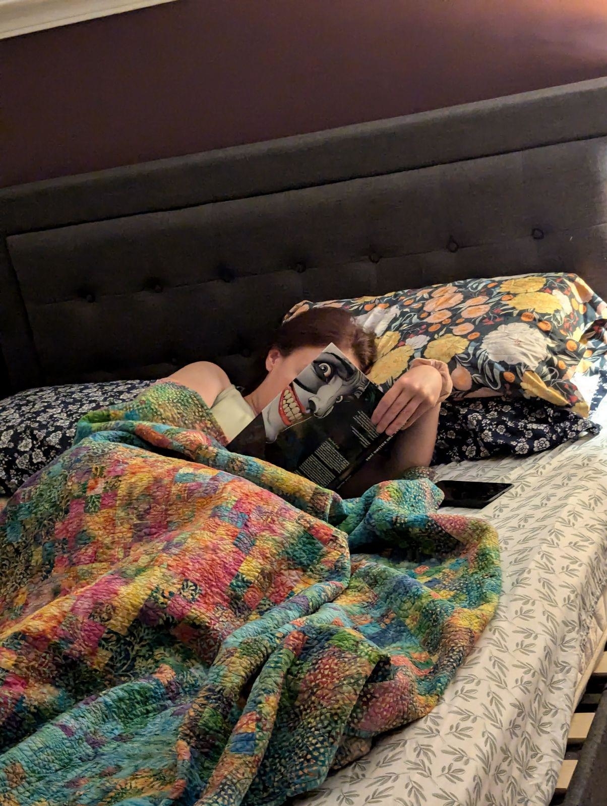 Person lying in bed under a colorful quilt, reading a book with a spooky cover featuring a grinning face