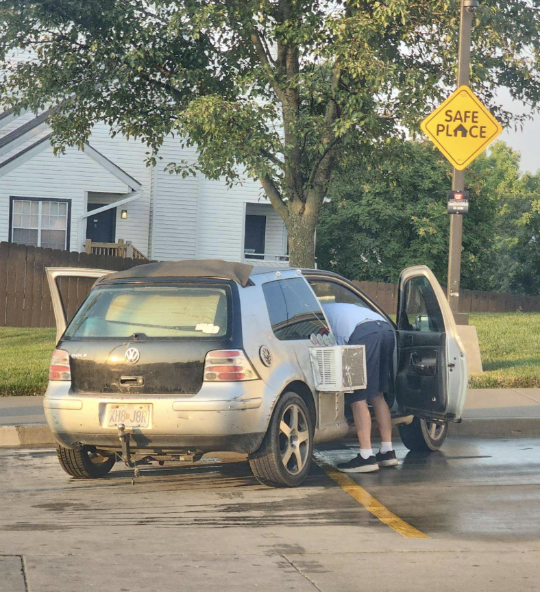 A person is bent over, looking inside the open door of a Volkswagon car in a parking lot. A &quot;Safe Place&quot; sign is visible on a light post nearby